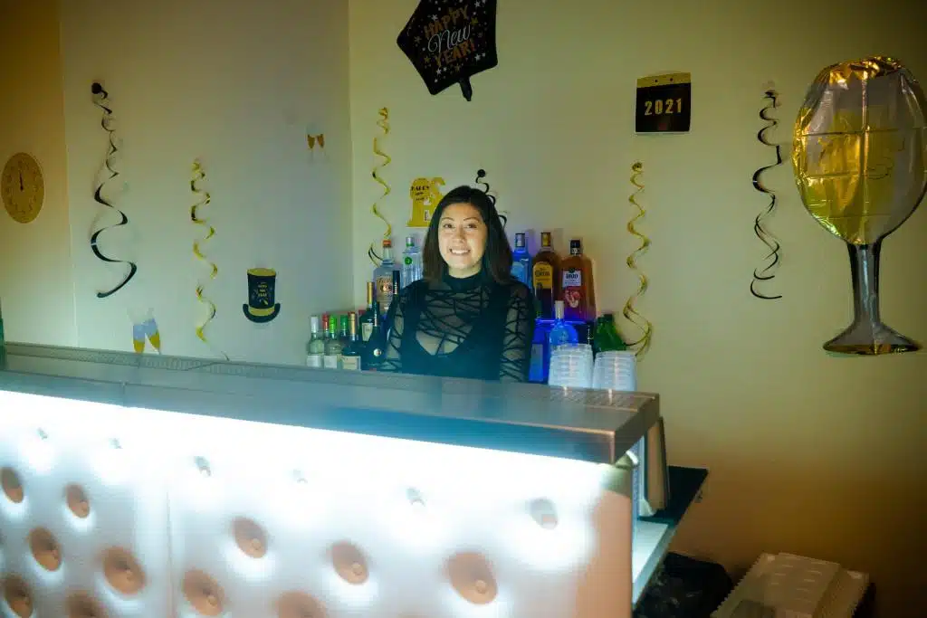 a female bartender behind white bar with bright lights. The bartender is smiling