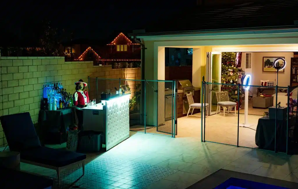 In backyard a white bar set up with LED lights. Inside of the bar a bartender with Christmas costume. On the right side of pictures, a photo booth set up.